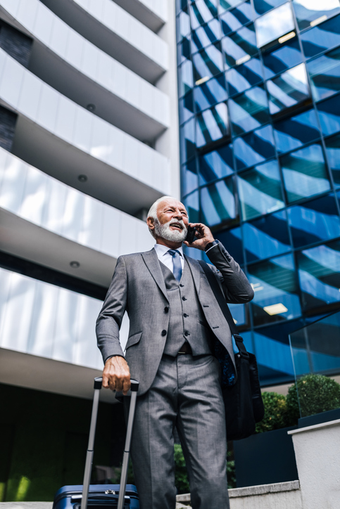 Smiling businessman with suitcase and bag talking on mobile phone during business travel.