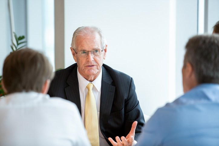 senior business man gesturing and talking to two partners who are sitting back to camera