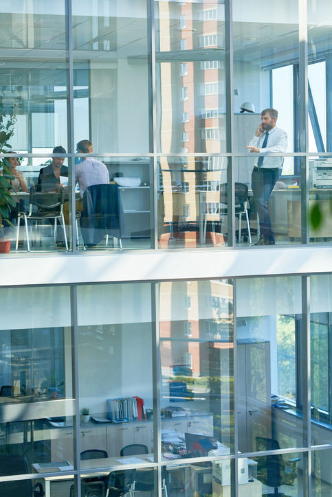 Wide angle portrait of mature businessman speaking by phone standing behind floor to ceiling windows in modern office building